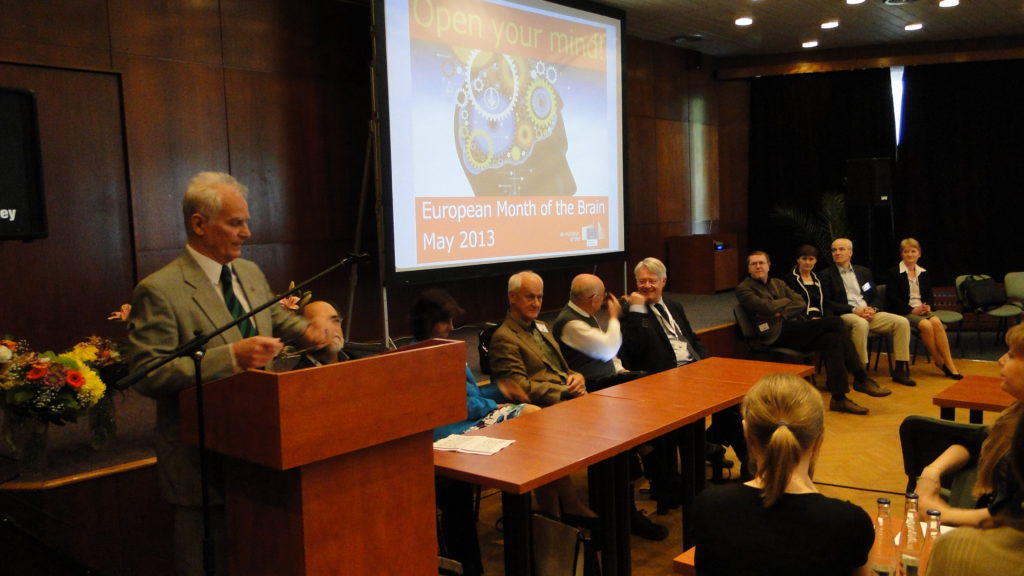 Man giving presentation at lectern. Screen showing slides in background, audience seated next and around lectern