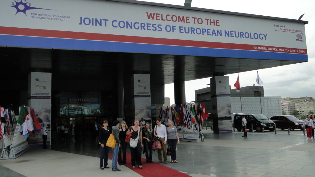 Seven people standing outside a conference venue under a sign that reads 'Welcome to the joint congress of European neurology'