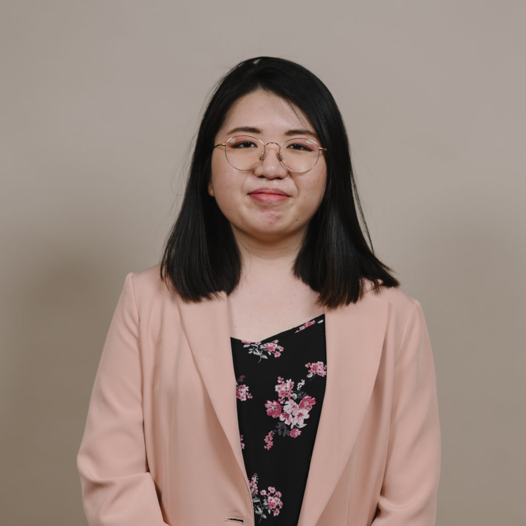 Chest height portrait of a young woman with shoulder length black hair, round glasses, and a peach-coloured blazer