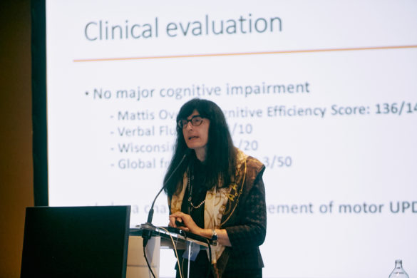 Prof. Elena Moro standing at lectern , speaking in front of a large screen displaying a slide from her presentation